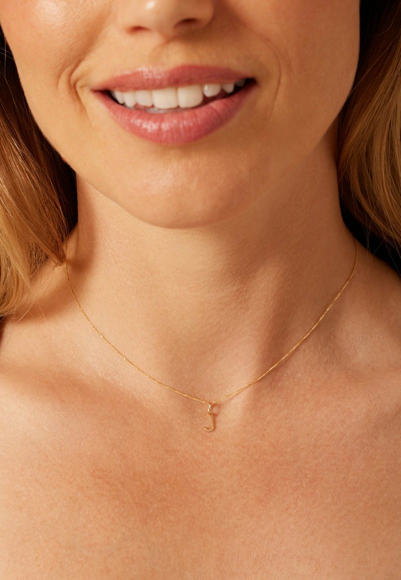 Collier en chaîne dorée avec un petit pendentif stylisé en forme de lettre "J". Design délicat, texture lisse, et maillons fins reposant sur une clavicule nue.