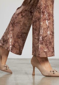Beige Wildleder High-Heels mit spitzem Zehenbereich und dekorativem Schleifendetail. Kombiniert mit weit geschnittenen, bedruckten, braunen Stoffhosen im Blumenmuster.