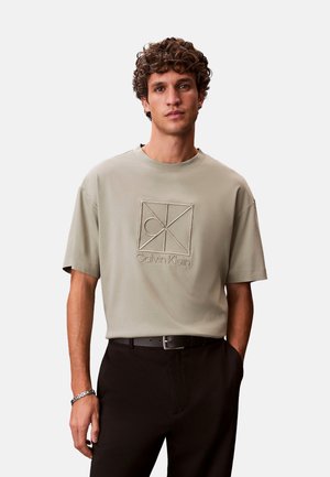 Joven con cabello rizado, vestido con una camiseta beige de Calvin Klein y pantalones negros, de pie con una mano en el bolsillo contra un fondo liso.