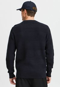 Navy strikkegenser med et teksturert chevronmønster, rund hals, ribbestrikkede mansjetter og kant. Stoffet ser mykt og tykt ut.