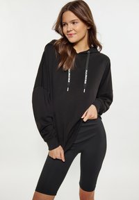 Felpa nera oversize con maniche lunghe, dotata di cappuccio regolabile e dettagli in metallo a contrasto, abbinata a leggings neri fino alla coscia.