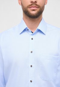 Camicia azzurro chiaro con bottoni, realizzata in tessuto testurizzato, con una tasca sul lato sinistro e bottoni marroni lungo la pattina.