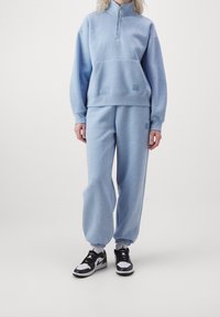 Helles blaues Sweatshirt mit Halb-Reißverschluss-Kragen und frontaler Tasche, kombiniert mit passenden Sweatpants. Beide sind weich, mit gerippten Bündchen und Logo-Akzenten.
