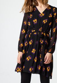 Robe noire avec de longues manches transparentes, col en V, et motif floral violet et jaune. Équipée d'une taille cintrée et d'une jupe fluide.