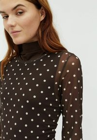 Brown, semi-sheer long-sleeve top with a high neckline, featuring a polka dot pattern in cream. Finished with a smooth trim.
