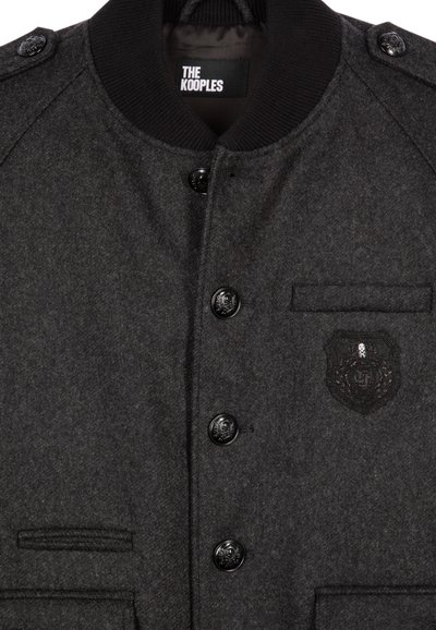 Grijze wollen blend jas met een geribde kraag, zwarte knopen, zijzak en een reliëfembleem op de borst. Het ontwerp is gestructureerd en klassiek.