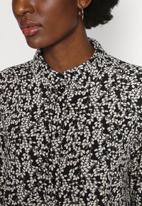 Blusa preta com padrão floral branco, tecido suave, gola clássica e fecho por botão. O padrão apresenta pequenas folhas e detalhes delicados.
