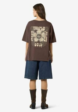 Femme portant un t-shirt marron oversize avec un grand motif de fleur et d'œil ainsi qu'un texte au dos, short en jean et bottes marron, vue de dos.