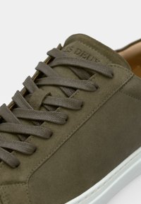 Olajzöld velúrbőr sneaker lapos, texturált fűzőkkel és fehér gumitalppal. Minimalista dizájn, diszkrét márkajelzéssel a nyelvén.