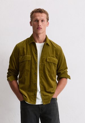 Olijfgroene corduroy knopenoverhemd met twee borstzakken, gedragen over een wit shirt. Model staat met handen in de zakken, grijze broek.