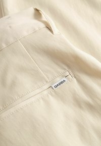 Tissu de couleur crème avec une texture côtelée fine, présentant une couture visible et une étiquette portant l'inscription "GABBA" cousue sur le côté.