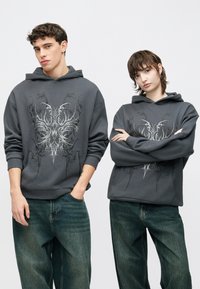 Šedá mikina s kapucňou s prepracovanou čiernou grafickou potlačou na prednej strane. Voľný strih s rebrovanými manžetami. Kombinovaná so svetlomodrými voľnými rifľami.