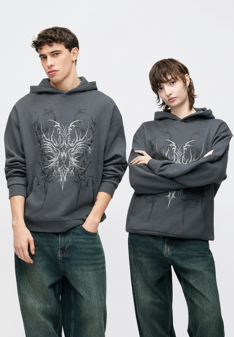 Šedá mikina s kapucňou s prepracovanou čiernou grafickou potlačou na prednej strane. Voľný strih s rebrovanými manžetami. Kombinovaná so svetlomodrými voľnými rifľami.