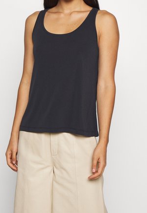 Femme portant un débardeur noir sans manches et un pantalon beige taille haute, montrée des épaules jusqu'à mi-cuisse sur un fond uni.