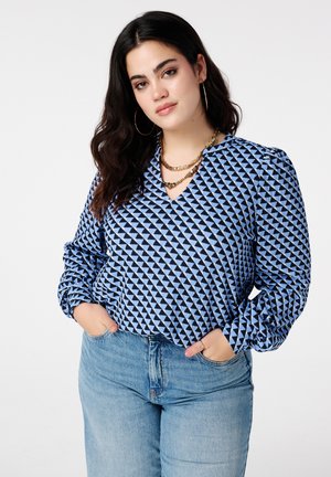 Vrouw met donker haar, grote ronde oorbellen, laagjes gouden kettingen, een blauw-zwarte blouse met geometrisch patroon en lichtblauwe spijkerbroek, handen in de zakken.