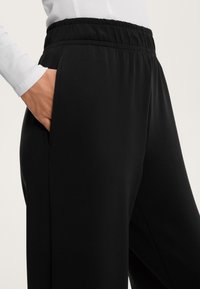 Svarta tapered byxor med elastisk midja och sidofickor. Slät textur med avslappnad passform, visad delvis med handen i en ficka.