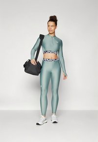 Türkizkék sportos crop top fél cipzárral és fekete logós derékszalaggal, hozzá illő leggingssel és fehér sportcipővel. A modell fekete táskát tart a kezében.