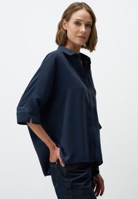 Navyblaues, oversized Hemd mit Knöpfen und Kragen sowie hochgeschlagenen Ärmeln; glattes Material, seitliche Schlitze und entspannter Schnitt. Kombiniert mit dunklen Jeans.