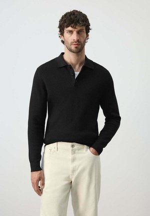Hombre con cabello rizado y oscuro, vestido con un polo negro de manga larga y pantalones beige claro, de pie con una mano en el bolsillo.