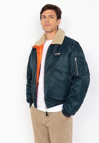 Marineblauwe bomberjack met een oranje voering en een beige nepbont kraag. Voorzien van ritsen en een logo-patch op de borst. Ribgebreide manchetten.