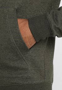 Handen inskjuten i framfickan på en mörkgrön prickig sweatshirt med ribbad mudd vid handleden.