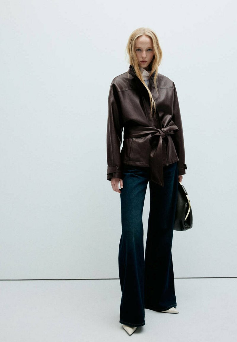 Femme portant une veste en cuir marron foncé ceinturée, un jean large foncé, des chaussures blanches pointues, tenant un sac à main noir sur un fond uni.