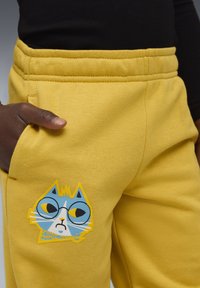 Gule sweatpants med en blå kattegrafik iført briller på frontlommen. Blødt, struktureret stof med elastisk talje.