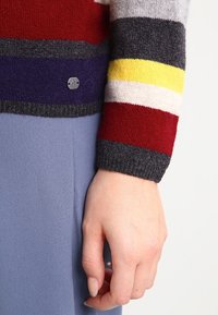Pull en tricot à rayures multicolores, comportant des rayures rouges, grises, jaunes et violettes, avec des poignets côtelés et un petit logo argenté.