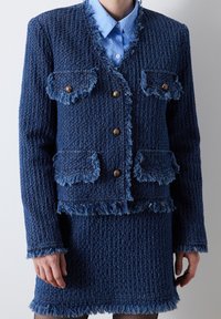 Ensemble en tweed bleu marine avec jacket et jupe, bord frangé, surface texturée, boutons dorés et poches plaquées. Chemisier bleu clair à col en dessous.