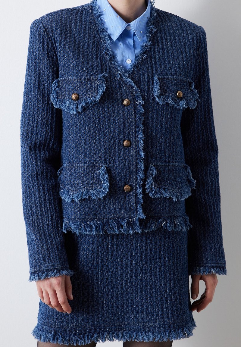 Ensemble en tweed bleu marine avec jacket et jupe, bord frangé, surface texturée, boutons dorés et poches plaquées. Chemisier bleu clair à col en dessous.