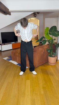 Balta svītraina īsām piedurknēm krekls, melnas bikses un baltas kurpes. Istaba ar koka grīdu, brūns dīvāns un zaļš augs podā.