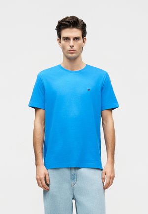 Fiatal férfi egyenesen áll, élénk kék rövid ujjú pólót és világoskék farmert visel, sima fehér háttér előtt.