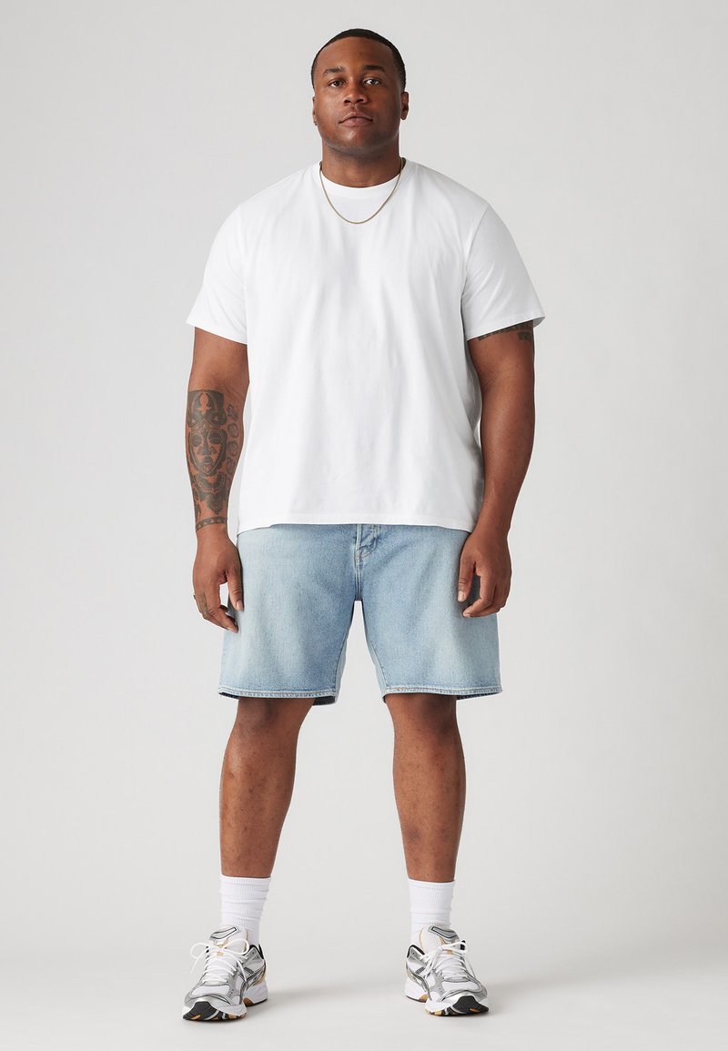 Homme avec un bras tatoué portant un t-shirt blanc, un short en denim clair, des chaussettes blanches, des baskets blanches et une fine chaîne en or, debout.