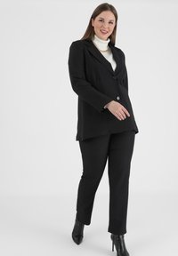 Sort skræddersyet blazer med en enkelt knap og taljerede revers, parret med matchende tætsiddende sorte bukser og spidse sorte hæle.