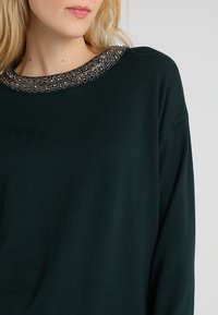 Maglione lavorato a maglia verde scuro con un ampio scollo decorato con perline intricate e accenti metallici lungo il colletto. Texture morbida.
