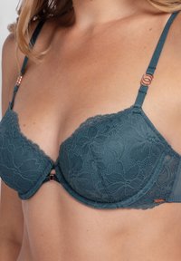 Blue lace bra with floral patterns and rose gold ring details on adjustable straps, worn by a person with light skin tone.