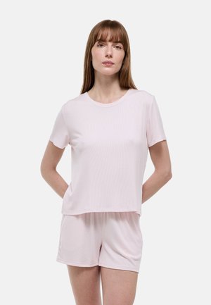 Femme aux cheveux bruns lisses portant un haut à manches courtes rose clair et un short assorti, debout avec les mains derrière le dos sur un fond blanc.