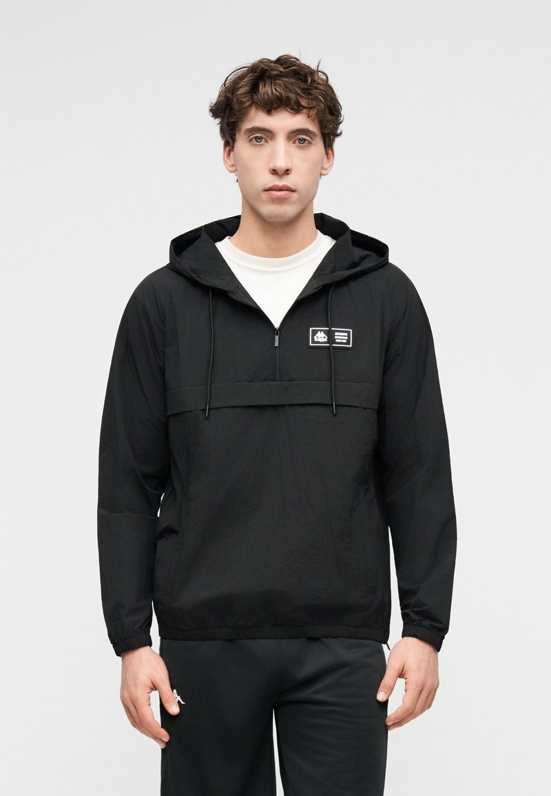 Νέος άνδρας που φοράει μαύρο ελαφρύ μπουφάν με κουκούλα και λευκή ετικέτα λογότυπου στο στήθος και μαύρο παντελόνι, σε απλό φόντο.