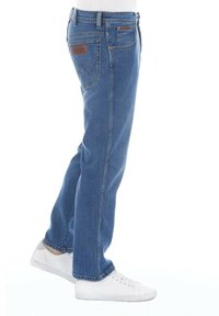 Jeans de mezclilla azul claro con corte recto, diseño de cinco bolsillos y parche de cuero marrón en la parte trasera de la cintura, combinados con zapatillas blancas.