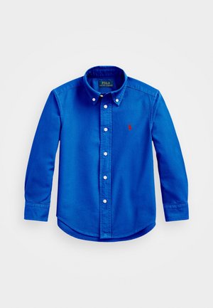 Niebieska koszula na długi rękaw z guzikami, kołnierzykiem zapinanym na guziki, białymi guzikami i czerwonym wyhaftowanym logo na piersi, ułożona płasko na szarym tle.