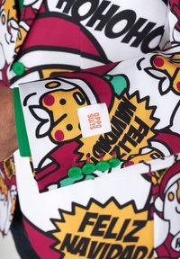 Heller Anzugjacke mit weihnachtlichen Motiven von Santa Claus und Urlaubssprüchen, aus einem Stoff mit glänzender Oberfläche. Grüne Knöpfe und sichtbares Markenetikett.
