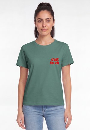 Frau mit dunklem, zurückgebundenem Haar, die ein grünes T-Shirt mit rotem Text "c'est la vie" und hellblaue Jeans vor weißem Hintergrund trägt.