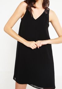 Svart ärmlös klänning med V-halsringning och spetsdetaljer på ena axeln. Tillverkad av lätt, flödande tyg med en mjuk textur.