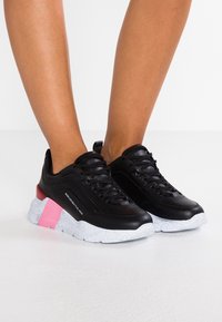 Baskets en cuir noir avec une finition texturée, présentant une semelle tachetée de blanc, des accents rose et rouge, et un design à lacets.