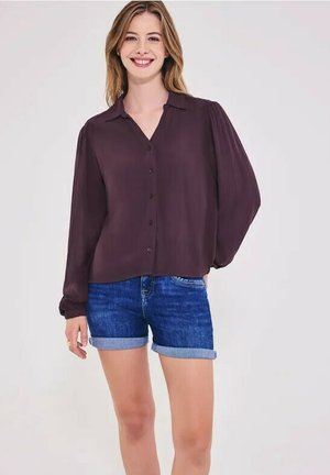 Junge Frau lächelt, trägt eine dunkelblaue Langarmbluse und hochgekrempelte Jeansshorts, steht vor einem einfarbigen hellen Hintergrund.