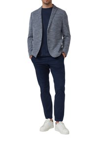 Grau strukturierter Blazer, marineblaues Hemd, marineblaue Hose und weiße Sneakers. Der Blazer hat einen Reverskragen und zwei Fronttaschen.