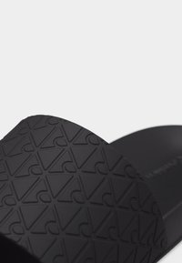 Sandale noire à glissière avec une semelle texturée présentant un motif géométrique répétitif de triangles et de demi-cercles.