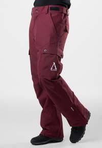 Bordowe wodoodporne spodnie cargo z bocznymi kieszeniami, logo na nodze, wzmocnione szwy i zamkiem błyskawicznym przy kostce; połączone z czarnym obuwiem.