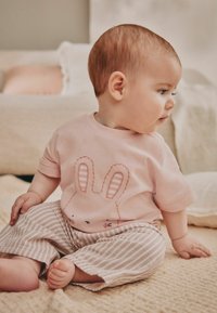 Camiseta de algodón rosa claro con diseño de conejo bordado, combinada con pantalones cortos a rayas beige, mostrando a un bebé sentado sobre una superficie texturizada.