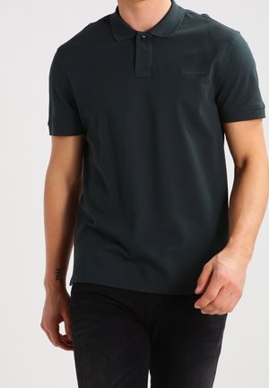 Man draagt een donkergroen poloshirt met korte mouwen en een zwarte broek, met een tatoeage op zijn linkervoorschouder.